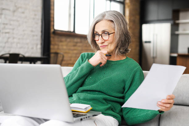 Serious senior accountant attentively looking at laptop screen, examines the budget, documents of the company, on the living room, holding laptop on lap, working online, prepares an annual report